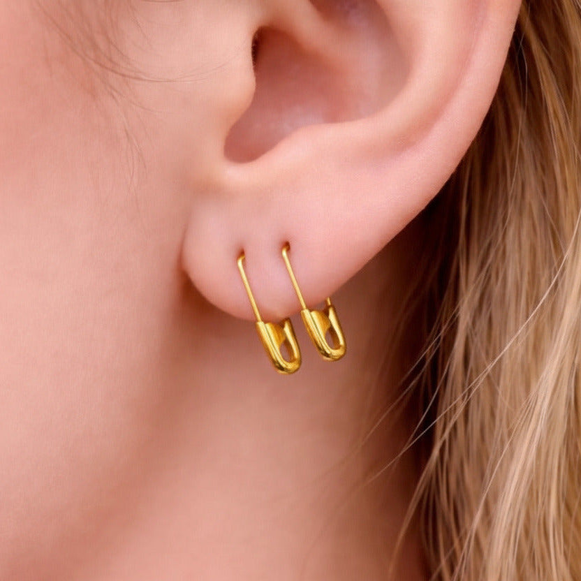 Small gold safety pin hoop earrings worn on pierced earlobe, minimalist punk style.