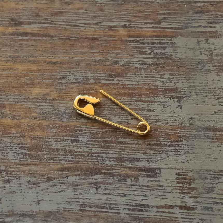 Small gold safety pin earring with shiny finish on rustic wooden surface.