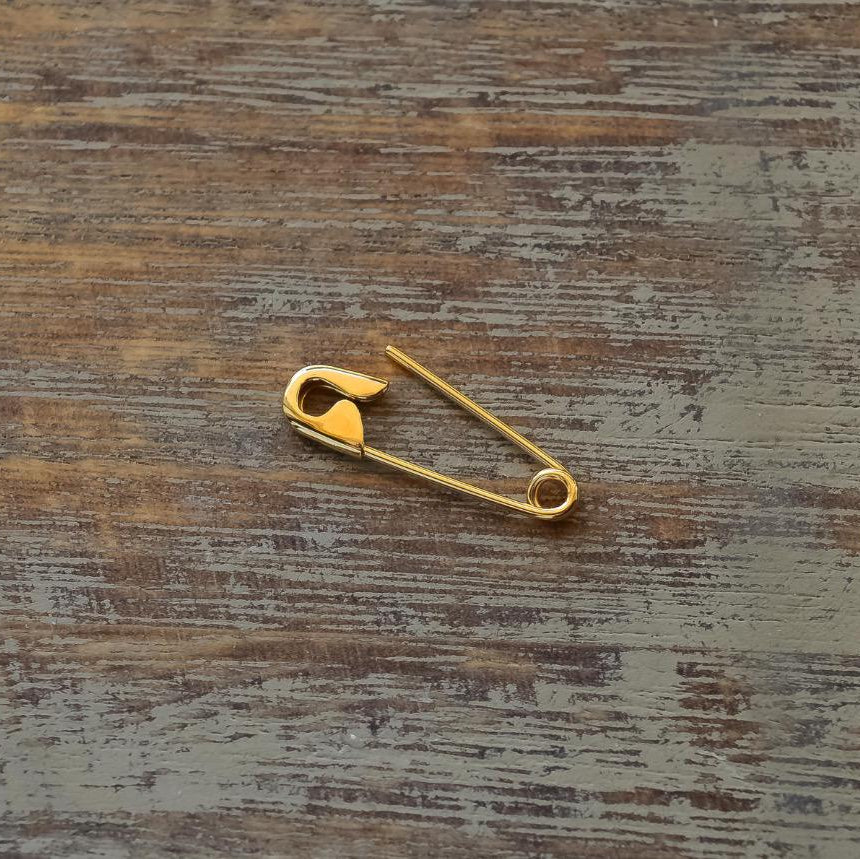 Small gold safety pin earring with shiny finish on rustic wooden surface.