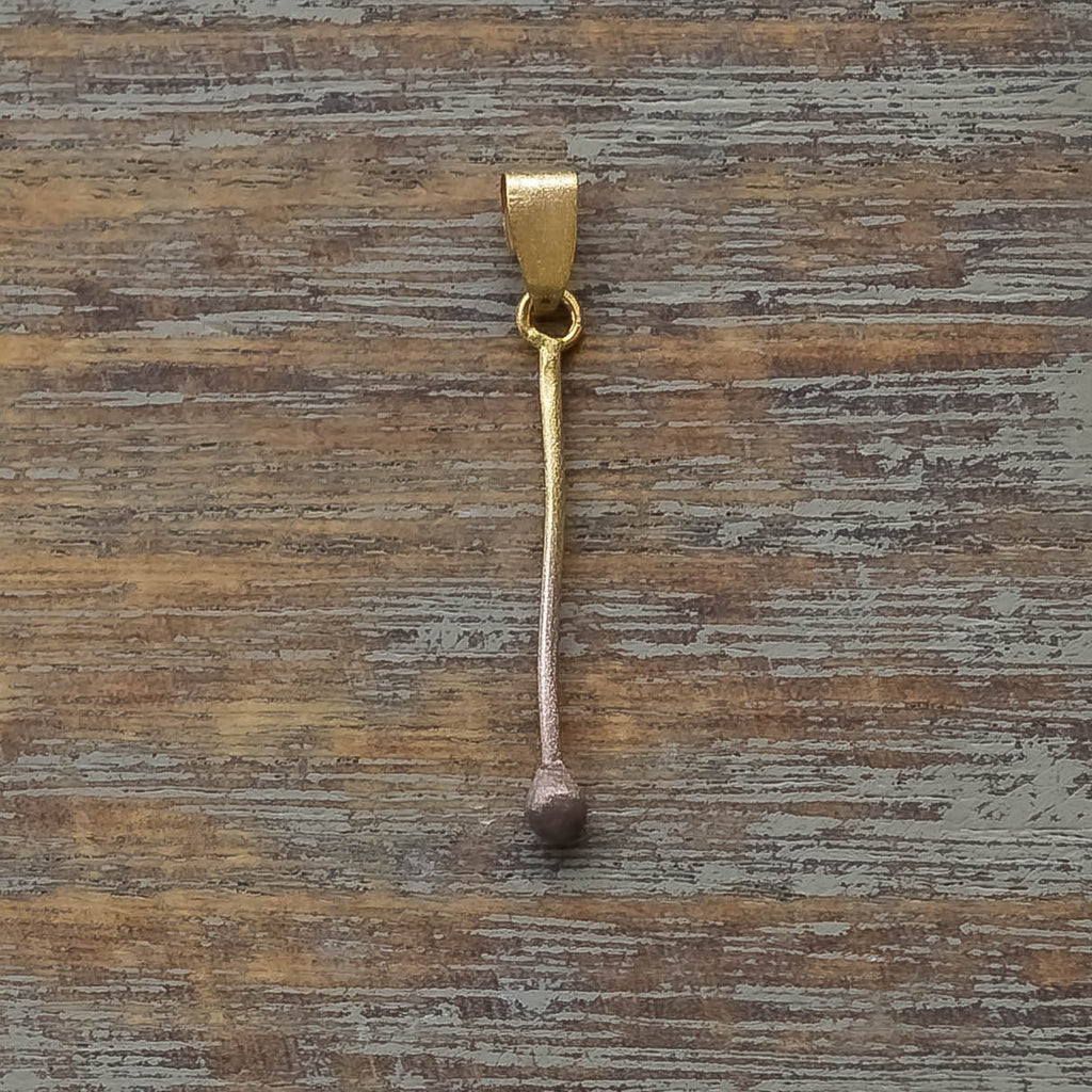 Minimalist gold pendant shaped like a slender matchstick with textured tip on wooden surface.