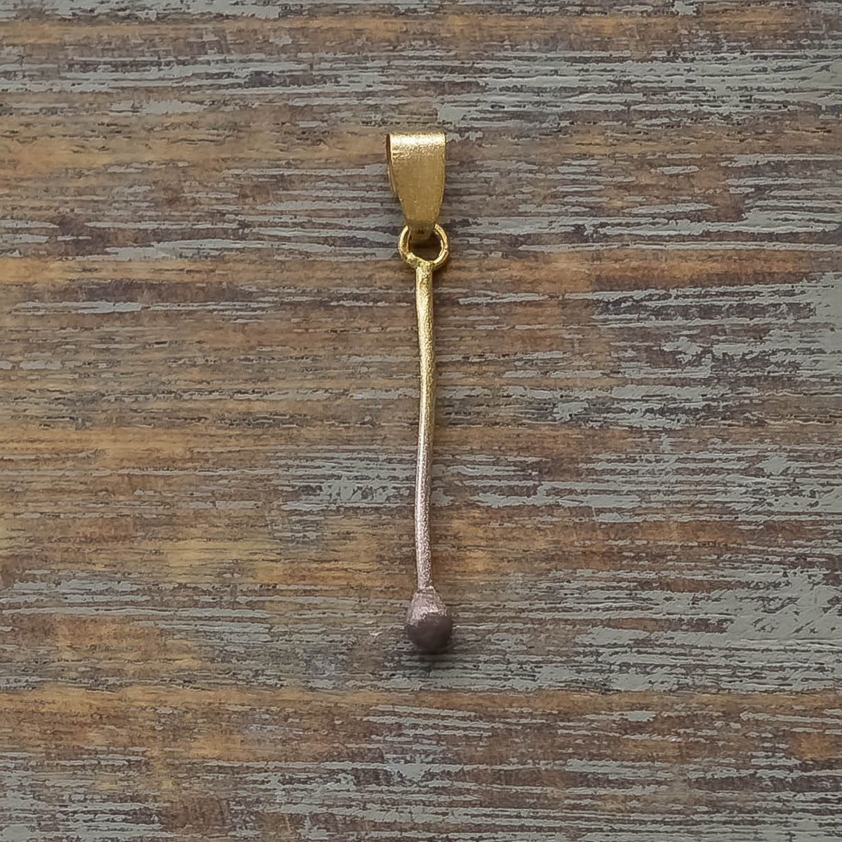 Minimalist gold pendant shaped like a slender matchstick with textured tip on wooden surface.