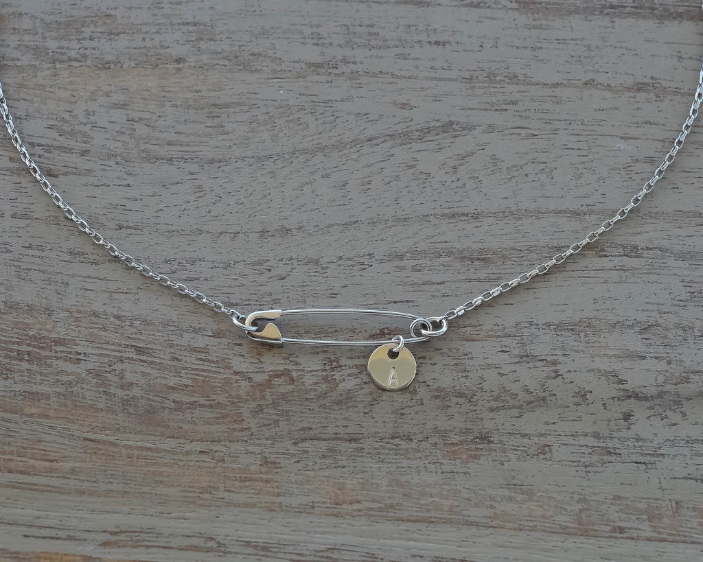Sterling silver safety pin necklace with small round initial charm on delicate chain on wooden surface .