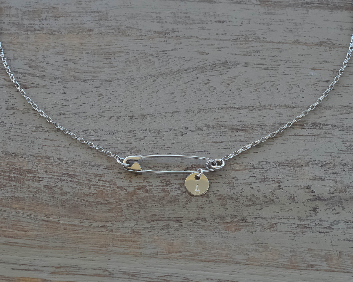 Sterling silver safety pin necklace with small round initial charm on delicate chain on wooden surface .