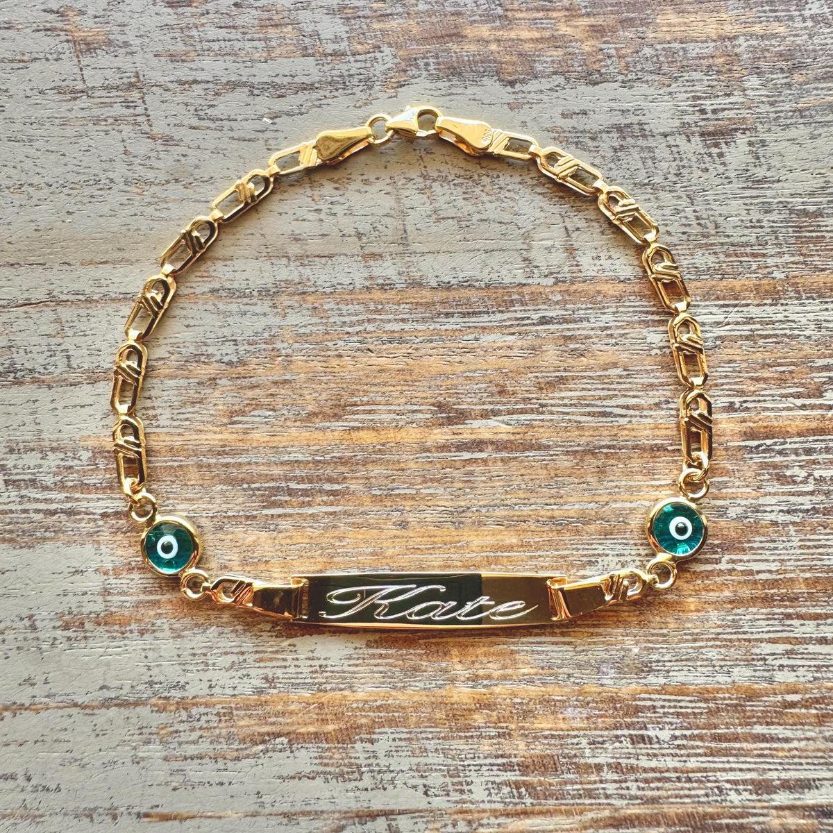 Personalized Baby Name Gold Bracelet – Nazar Protection Charms & Cursive Engraving on wooden surface - 24jewels