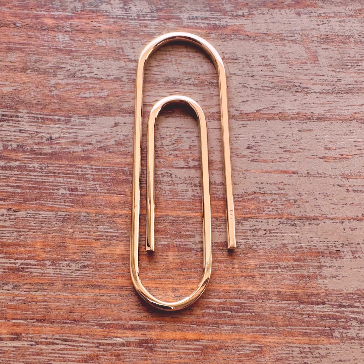 Handcrafted rose gold metal paperclip-shaped money clip with a smooth polished finish on wood surface.