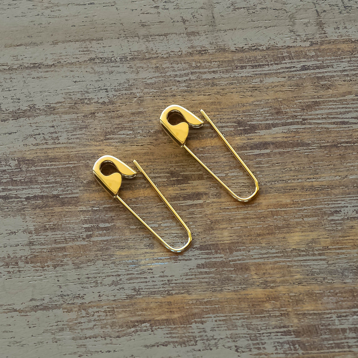 Pair of gold plated safety pin silver earrings with a shiny finish on a rustic wooden surface.
