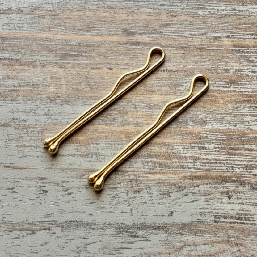 Two sterling silver bobby pins gold plated with a minimalist design, rounded ends, and a smooth metallic finish on a wooden surface.