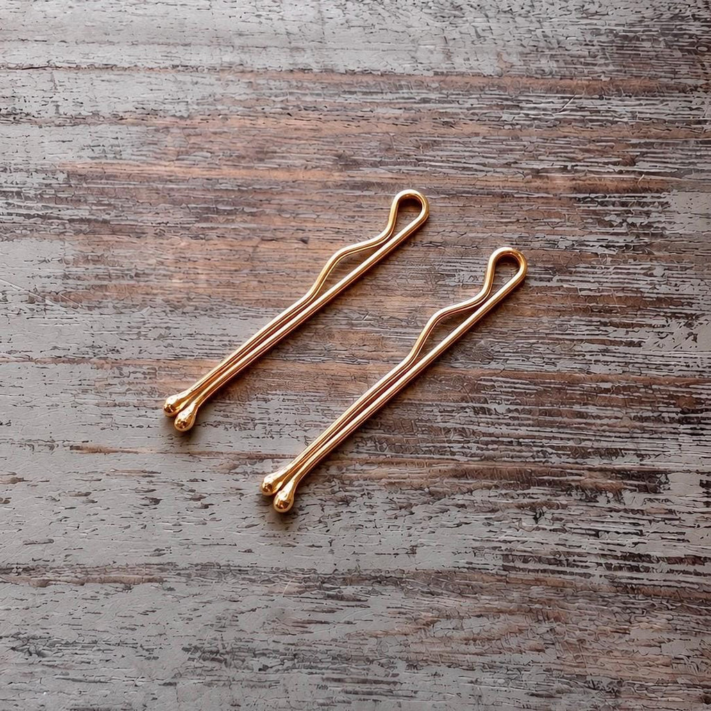 Two slender sterling silver bobby pins rose gold plated with a smooth, polished finish resting on a rustic wooden surface.
