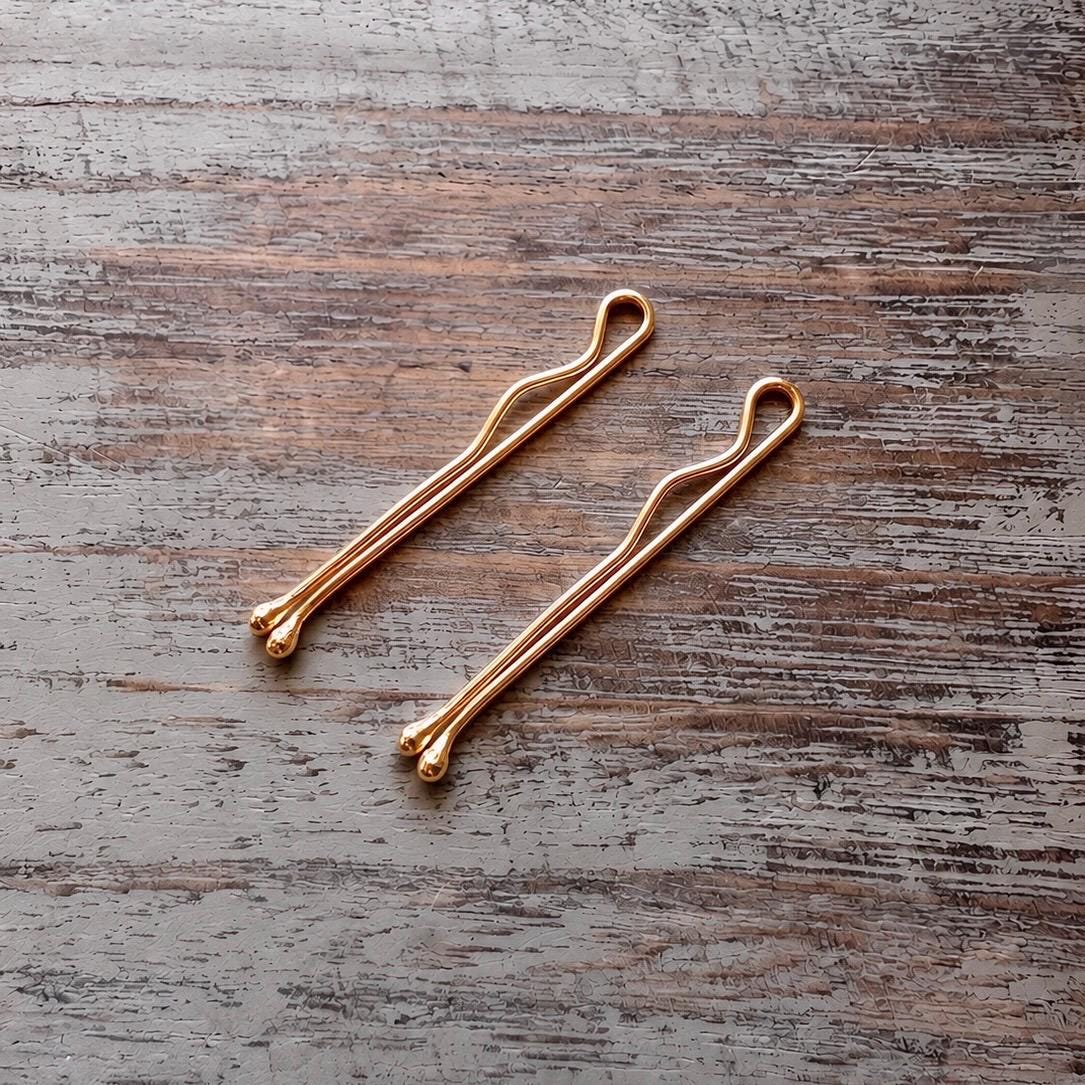 Two slender sterling silver bobby pins rose gold plated with a smooth, polished finish resting on a rustic wooden surface.