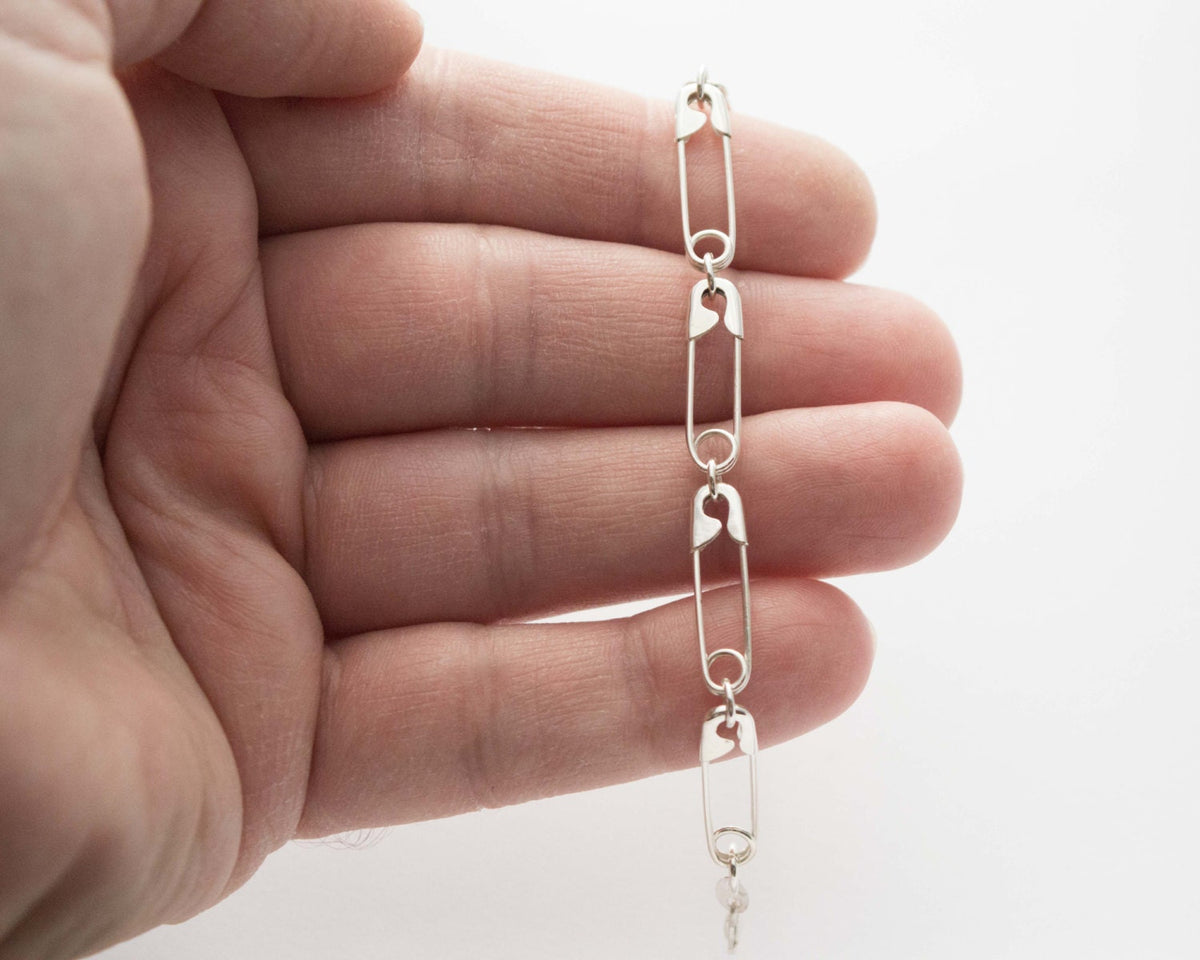 Sterling silver chain necklace with linked safety pin-shaped charms held between fingers against a white background.