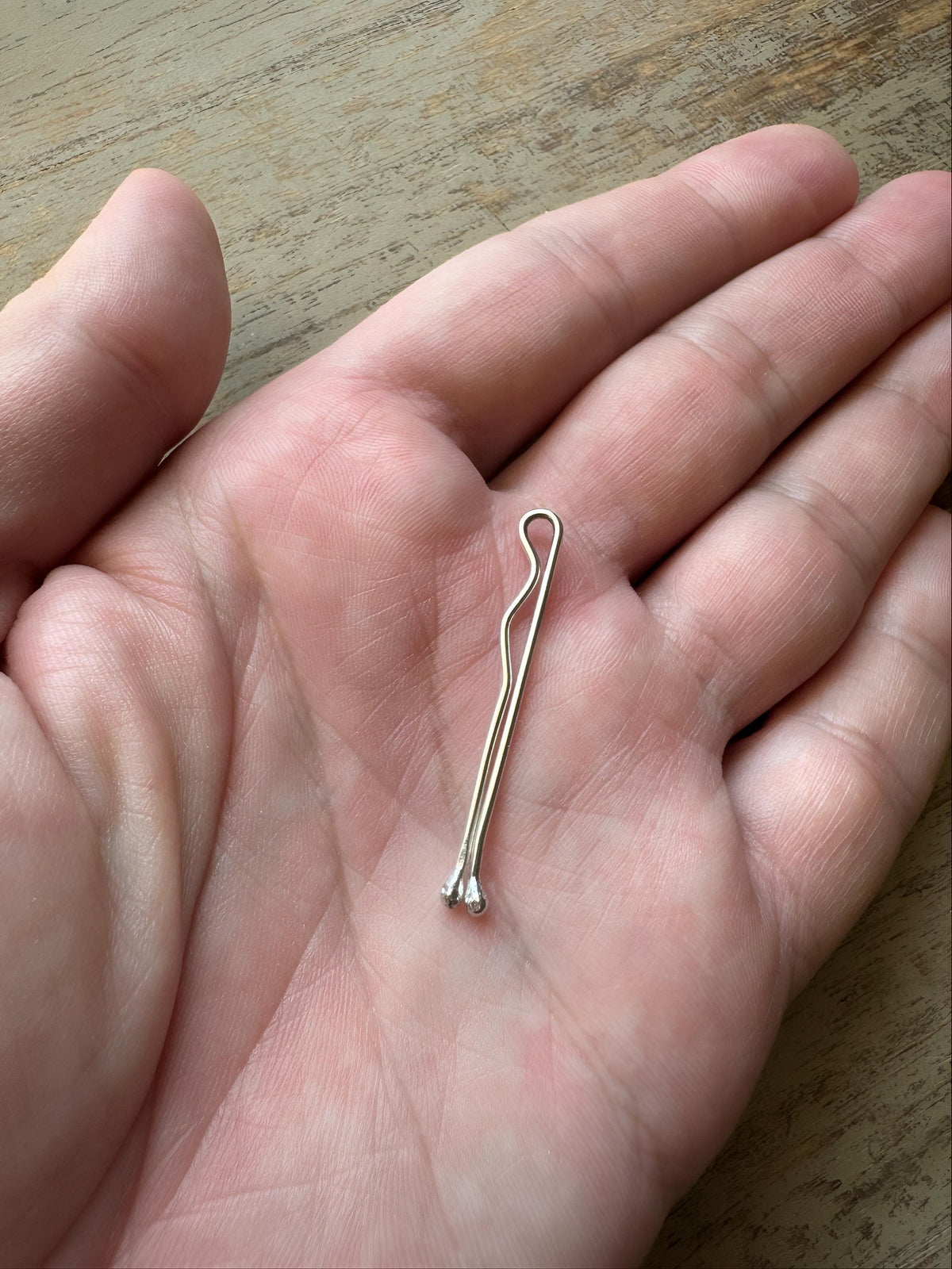 Small slender gold bobby pin with rounded tip, held in an open palm  of a man on wooden surface.