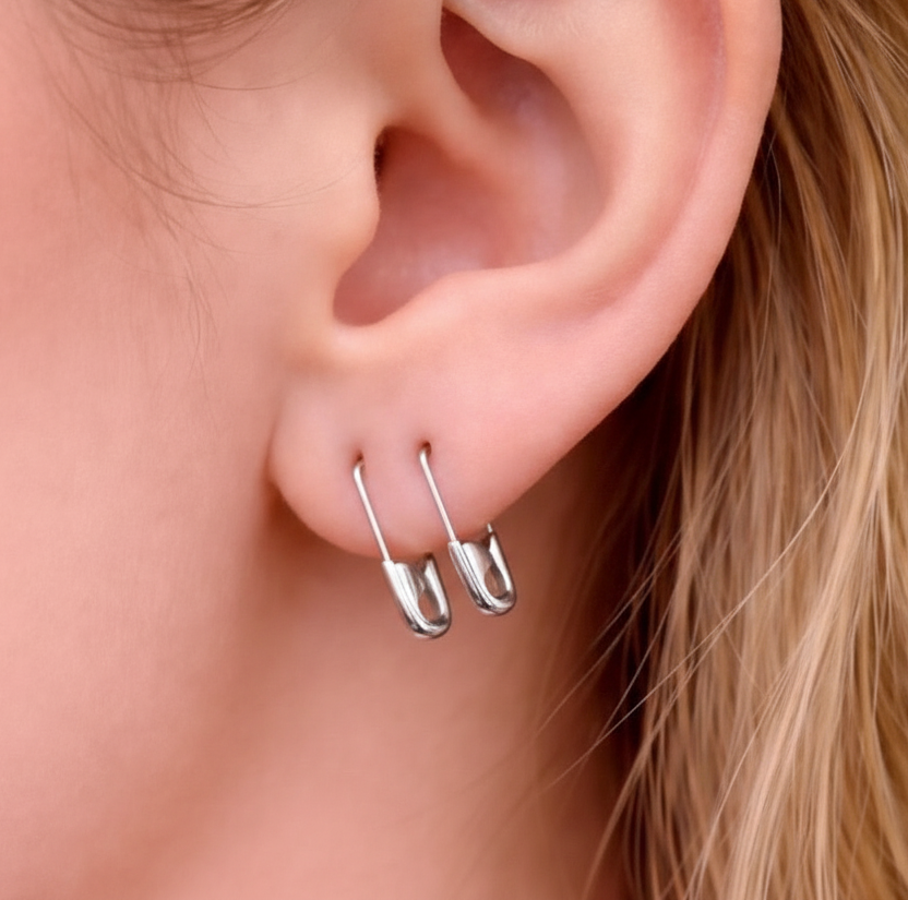 Small white gold hoop earrings shaped like safety pins with a shiny finish worn on earlobe .