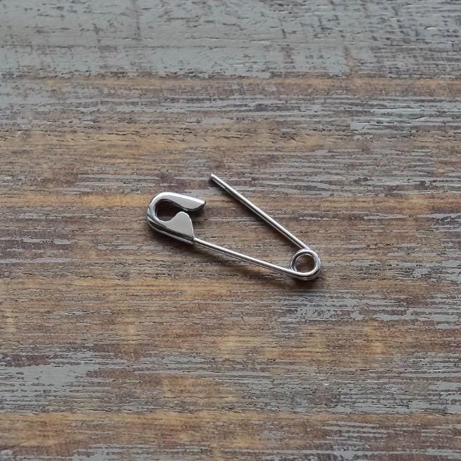 Small white gold safety pin earring with smooth, polished finish on rustic wooden surface.