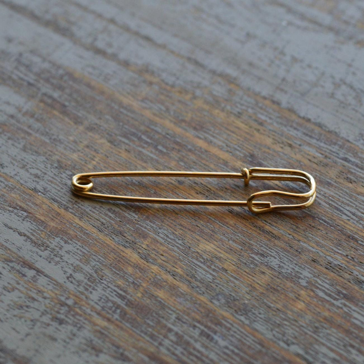 Delicate 1.25-inch yellow gold wire safety pin earring on wooden surface.