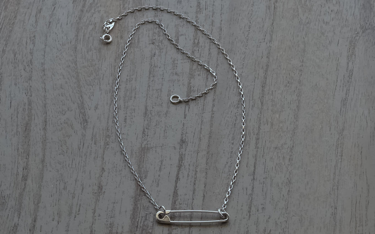 Sterling silver chain necklace with a minimalist safety pin charm on a wood surface.